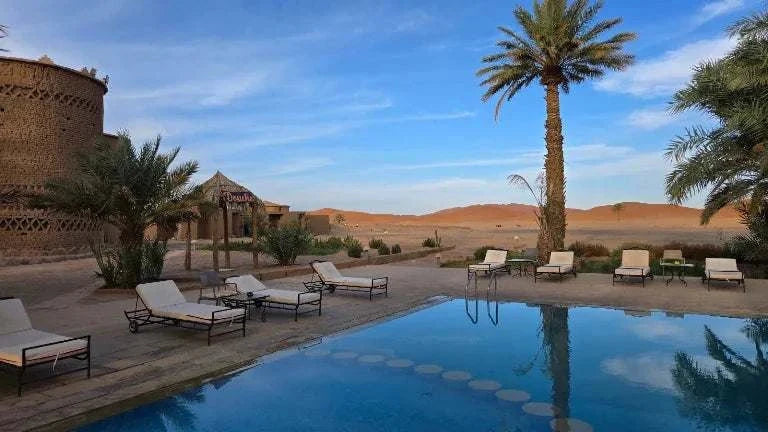 Kasbah Hotel Tombouctou outdoor pool area with lounge chairs, palm trees, and desert dunes in the background under blue sky