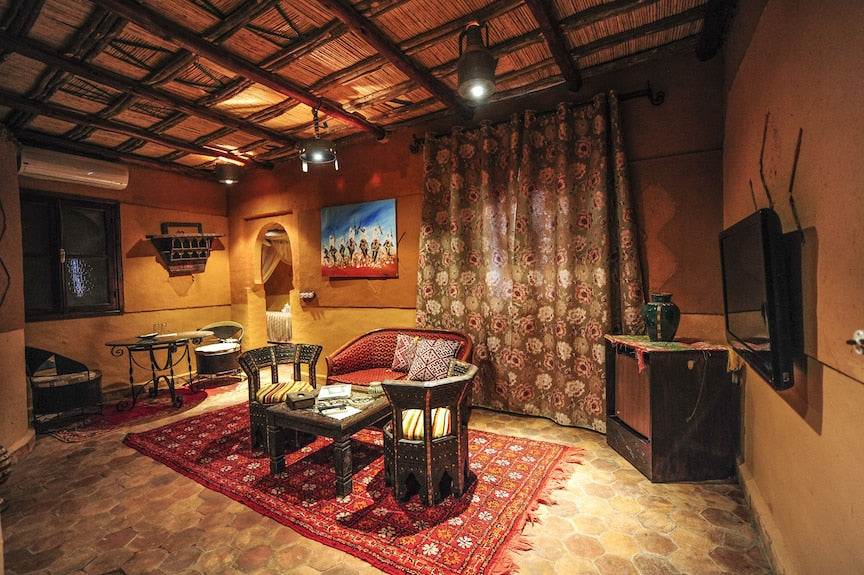 Kasbah Hotel Xaluca living room with warm adobe walls, rustic wood ceiling, patterned red rug, and wicker furniture with cushions