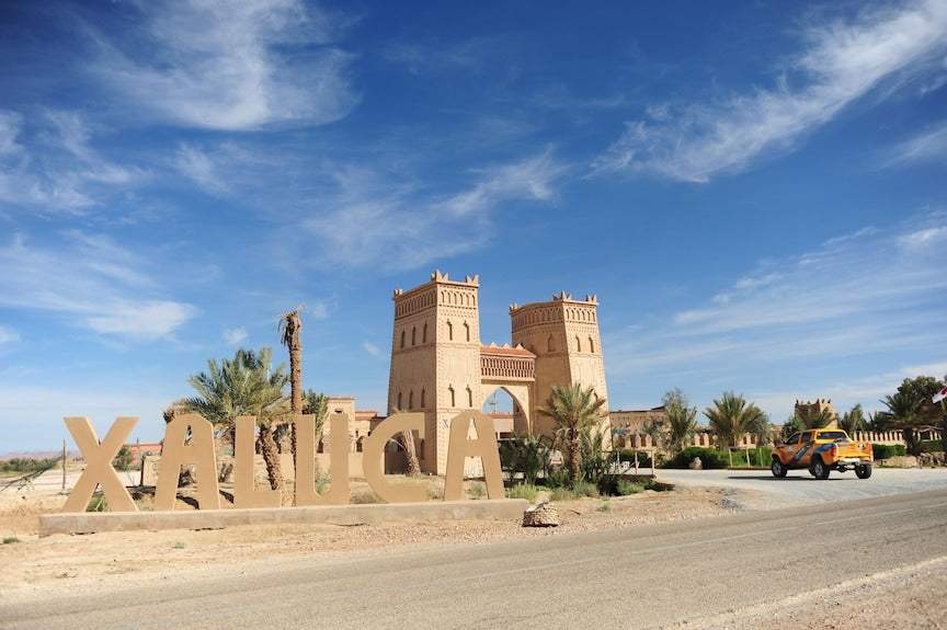 Kasbah Hotel Xaluca with large beige adobe towers and archway under a blue sky with scattered clouds and an XALUCA sign