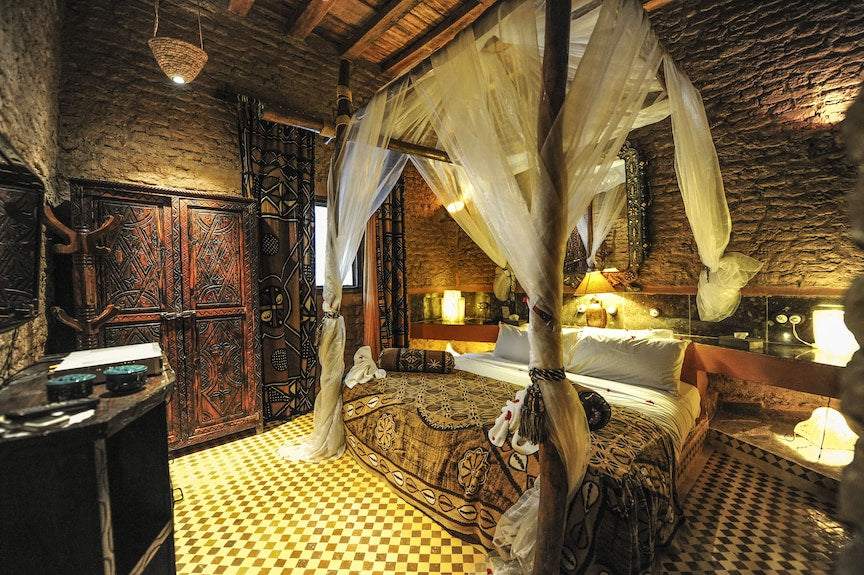 Kasbah Hotel Xaluca bedroom with earthy adobe walls, wooden four-poster bed draped in sheer fabric, patterned bedding, and rustic decor