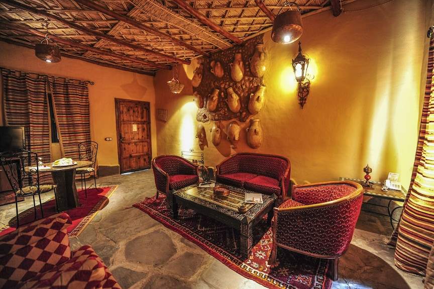 Kasbah Hotel Xaluca cozy sitting area with red patterned upholstery, traditional Moroccan rugs, clay pots wall decor, and warm lighting