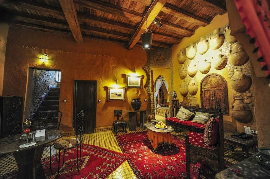 Kasbah Hotel Xaluca living room with warm orange walls, decorative plates, wooden ceiling, red patterned rugs, and traditional Moroccan furniture