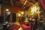 Kasbah Hotel Xaluca living room with warm orange walls, decorative plates, wooden ceiling, red patterned rugs, and traditional Moroccan furniture