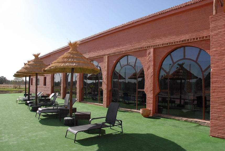 Kasbah Hotel Xaluca with terracotta adobe exterior walls, large arched windows, and straw parasols over black lounge chairs on green artificial grass