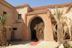 Kasbah Hotel Xaluca entrance with traditional adobe walls, ornate wooden door, and surrounding palm trees under clear sky