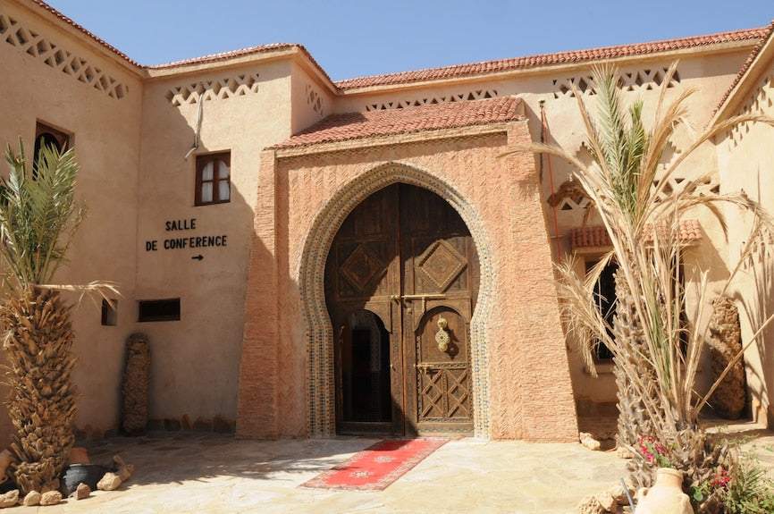 Kasbah Hotel Xaluca entrance with traditional adobe walls, ornate wooden door, and surrounding palm trees under clear sky