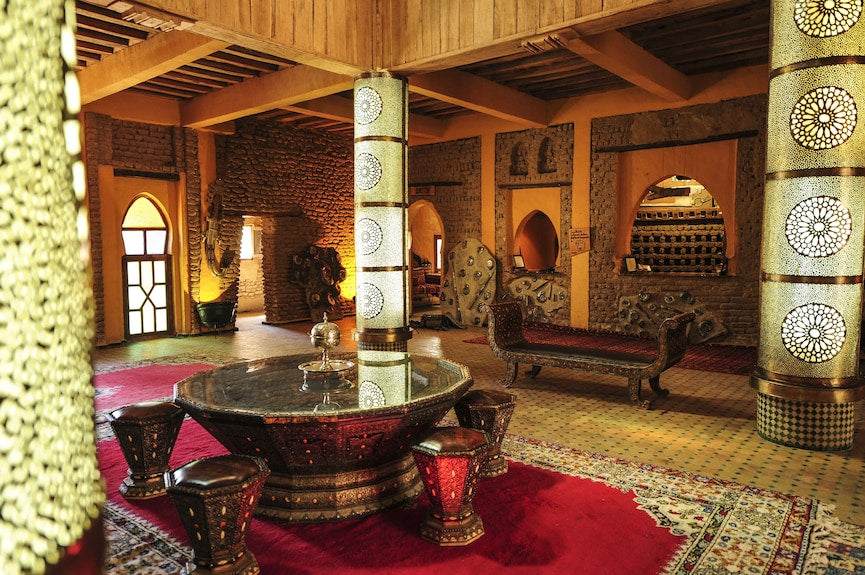 Kasbah Hotel Xaluca interior with traditional Moroccan wooden furniture, decorative pillars, intricate patterned carpets, and adobe walls