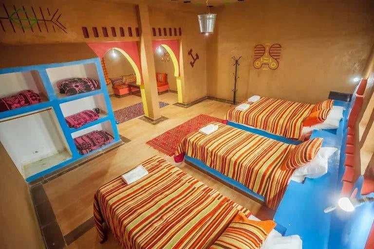 Kasbah Le Touareg traditional Moroccan-style bedroom with three single beds featuring striped orange and red bedding, bright blue furniture, and colorful wall decorations
