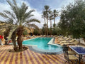 Outdoor swimming pool at Kasbah Le Touareg surrounded by palm trees, beige tiled deck, sun loungers, and a metal table with chairs.