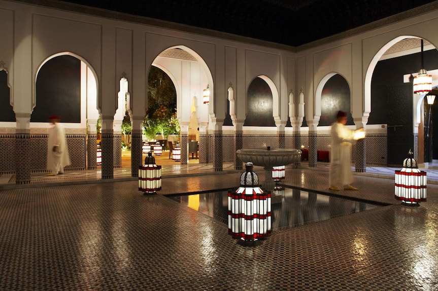 La Mamounia Marrakech Moroccan courtyard with intricate mosaic tiles, arches, lanterns, and blurred figures in traditional attire at night