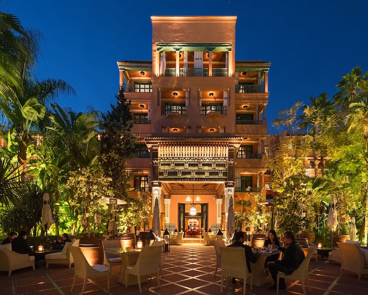 La Mamounia Marrakech luxury hotel exterior at night with warm lighting, Moroccan architecture, and outdoor dining area surrounded by lush greenery