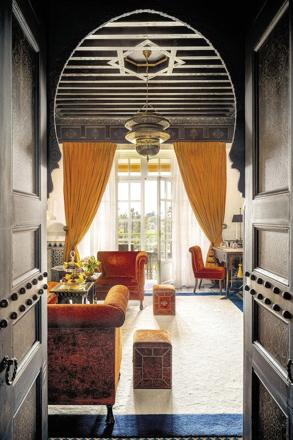 La Mamounia Marrakech living room with rich orange velvet chairs, intricate wooden arch doorway, Moroccan lantern, and bright natural light.