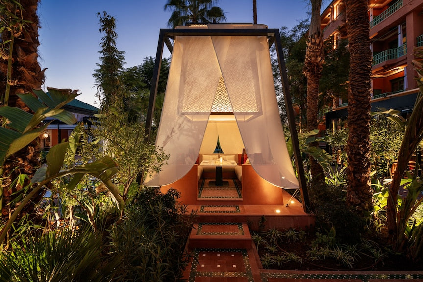 La Mamounia Marrakech outdoor luxury seating area with flowing white curtains, Moroccan tile flooring, and lush garden surroundings in warm evening light