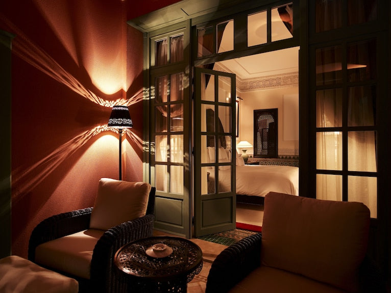 La Mamounia Marrakech outdoor seating area with dark wicker chairs, beige cushions, intricate metal table, and warm wall lamp casting shadows