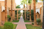 Le Berbere Palace Ouarzazate featuring beige stucco walls, palm trees, and a tiled walkway through a courtyard area