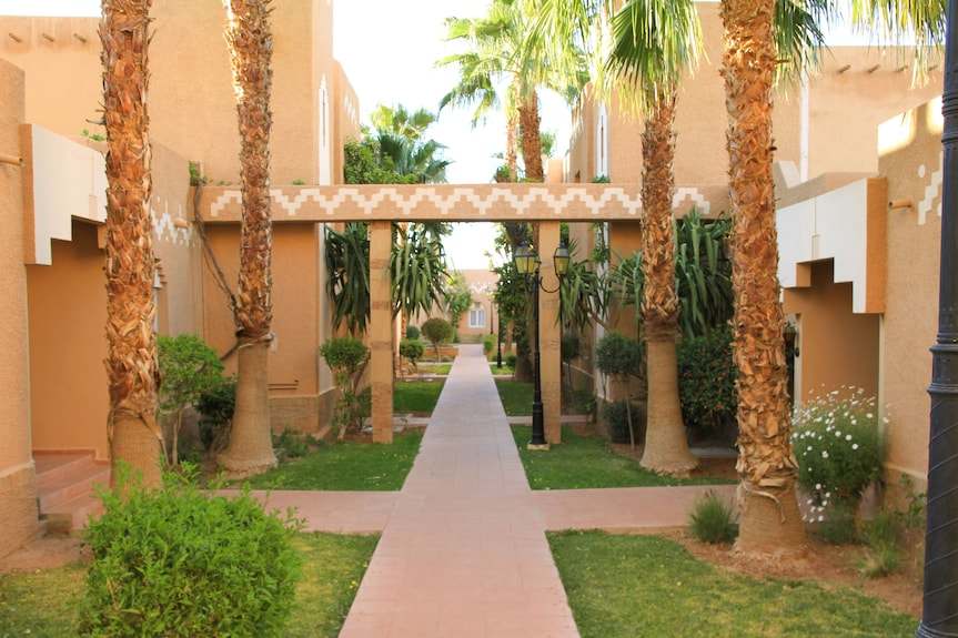 Le Berbere Palace Ouarzazate featuring beige stucco walls, palm trees, and a tiled walkway through a courtyard area