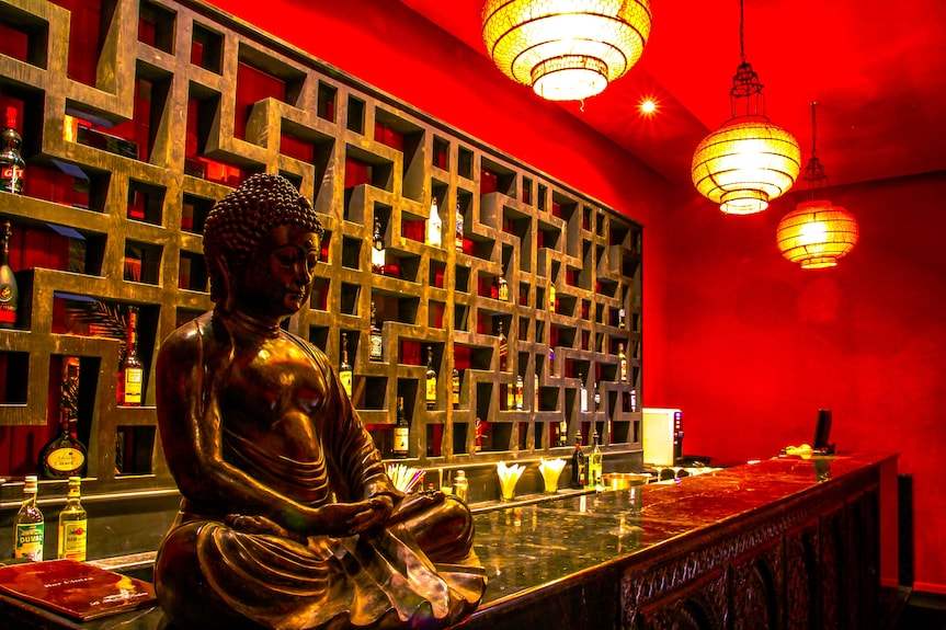 Le Berbere Palace Ouarzazate bar area with bronze Buddha statue, red walls, decorative shelving, and hanging lanterns