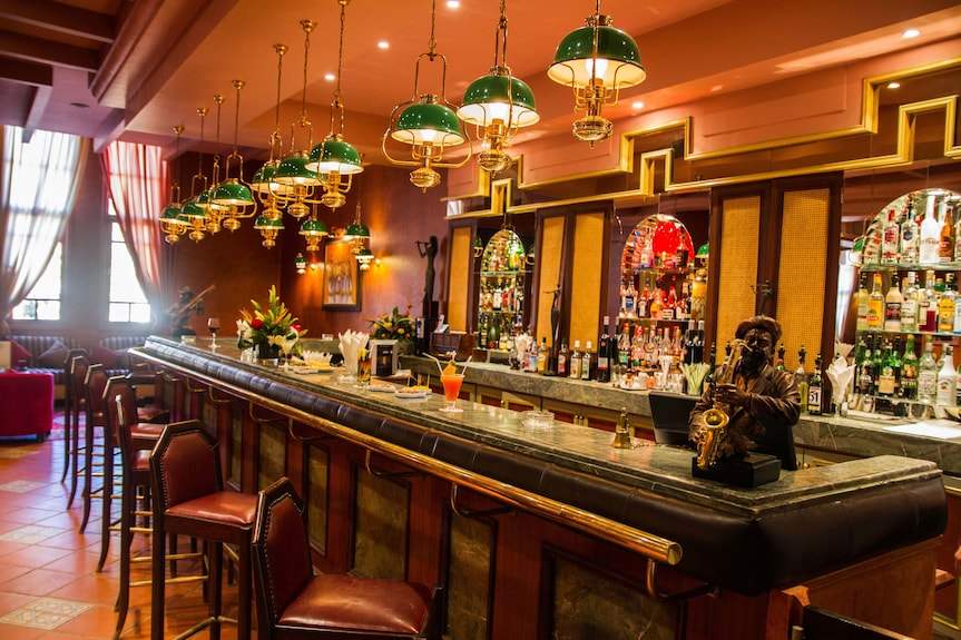 Hotel bar area with wooden paneling, leather chairs, green hanging lamps, and decorative statue at Le Berbere Palace Ouarzazate