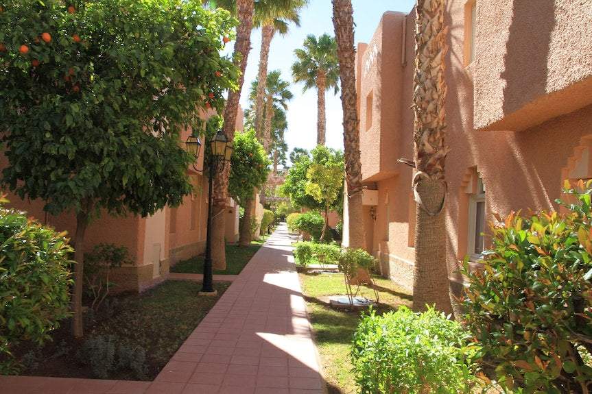 Le Berbere Palace Ouarzazate hotel outdoor walkway with terracotta walls, green palm trees, and shrubs under clear sky