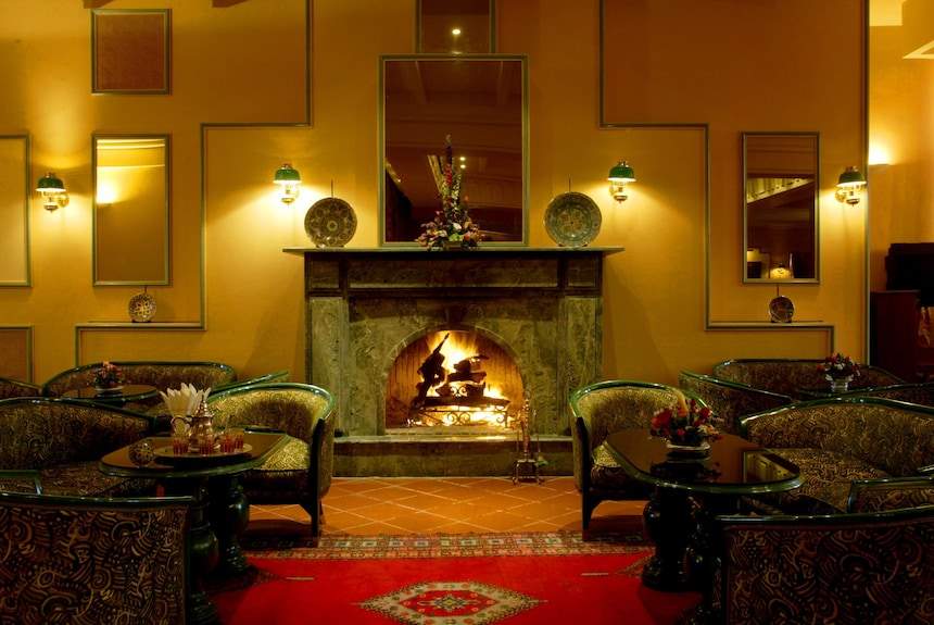 Le Berbere Palace Ouarzazate warm indoor lounge area with patterned armchairs, a stone fireplace, and a red rug