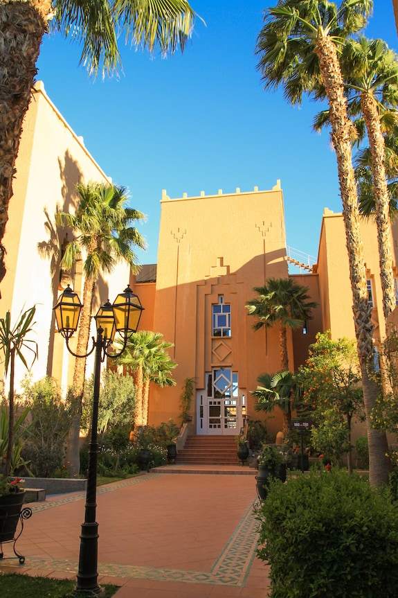 Le Berbere Palace Ouarzazate luxury hotel in Morocco with peach-colored towers, lush palm trees, and decorative entrance pathway