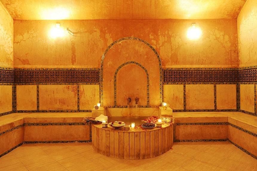 Le Berbere Palace Ouarzazate spa room with warm orange marble walls, traditional Moroccan tile accents, and a central wooden platform with towels and candles