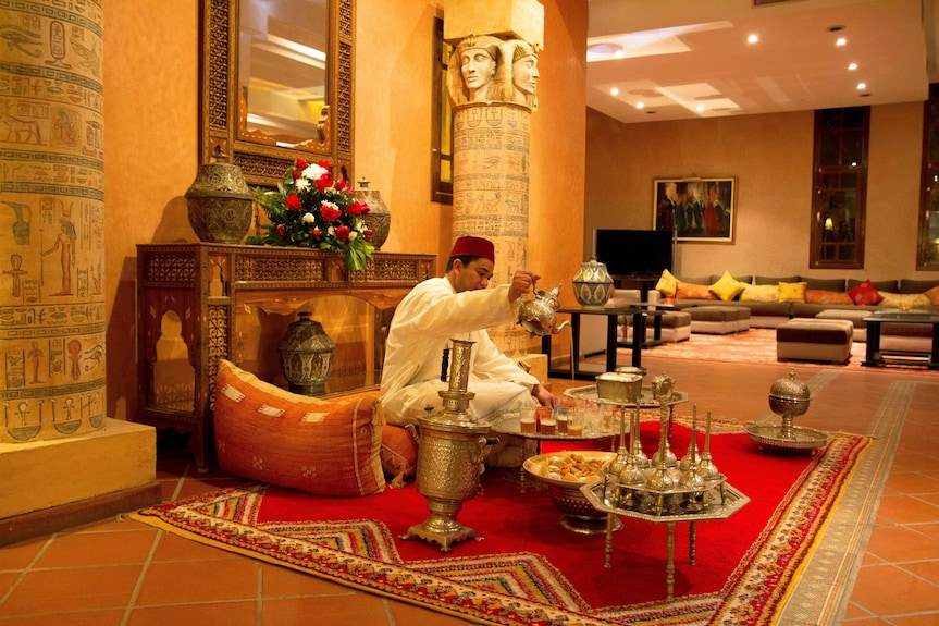 Le Berbere Palace Ouarzazate traditional Moroccan lounge with ornate silver tea set, red carpet, orange cushions, and intricate decor