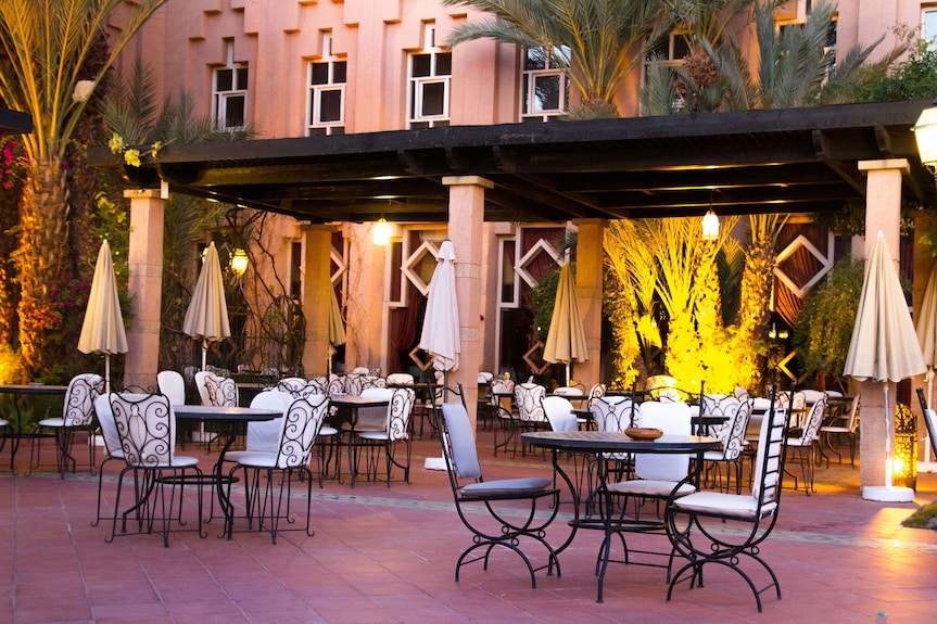 Le Berbere Palace Ouarzazate outdoor dining area with wrought iron chairs, round tables, beige umbrellas and warm lighting