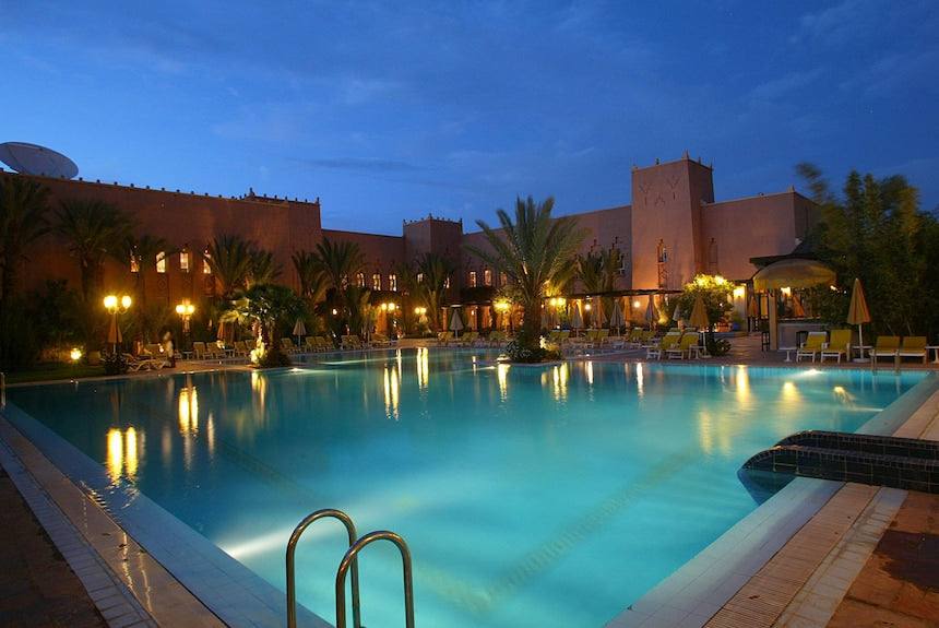Le Berbere Palace Ouarzazate outdoor swimming pool at dusk with illuminated water, surrounding lounge chairs and palm trees