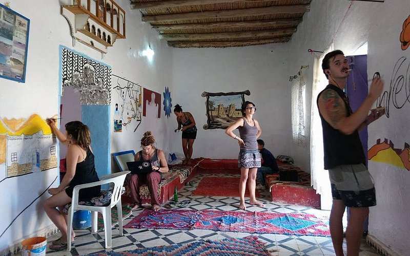 Le Gout Du Sahara Hostel interior with white walls decorated with murals, wooden ceiling, tiled floor, and colorful rugs, people painting and relaxing.