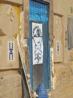 Blue wooden door with a white graffiti-style drawing on a textured tan wall, highlighting rustic charm at Le Gout Du Sahara Hostel.