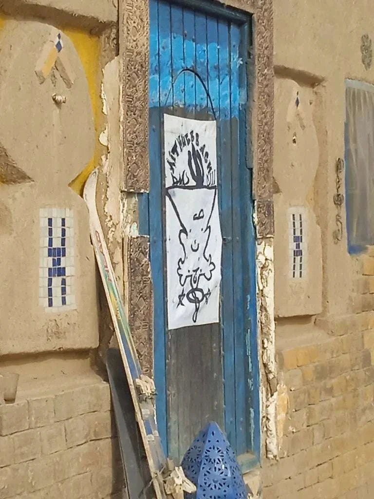 Blue wooden door with a white graffiti-style drawing on a textured tan wall, highlighting rustic charm at Le Gout Du Sahara Hostel.