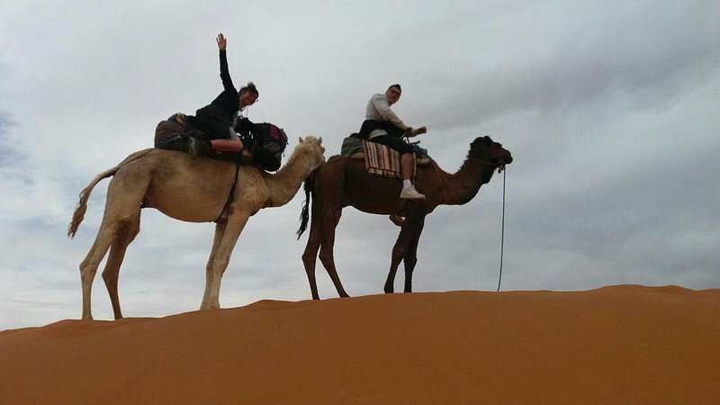 Two camels with riders on a golden sand dune under a cloudy sky, showcasing an adventurous experience at Le Gout Du Sahara Hostel.