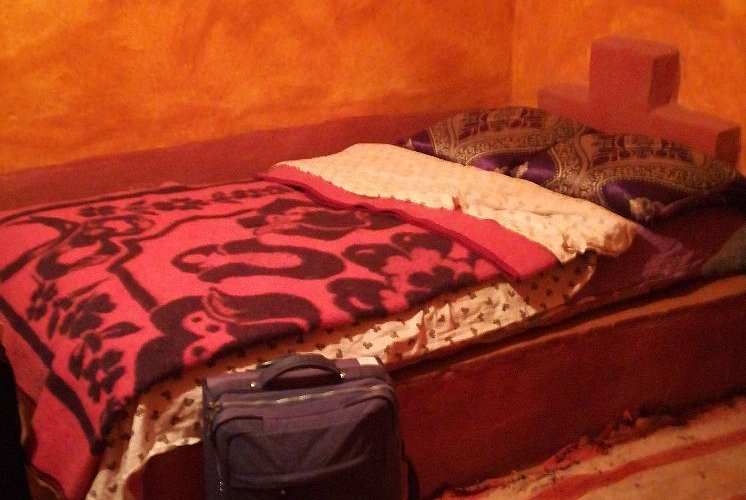 Single bed with patterned red and black blanket, purple decorative pillow, and suitcase on floor in a rustic room at Le Gout Du Sahara Hostel.