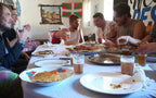 Group of people enjoying casual meal with flatbreads and glasses of tea around a white tablecloth, showcasing communal dining at Le Gout Du Sahara Hostel.