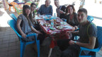 Group of five people sitting on blue plastic chairs around a table with red patterned cloth, sharing a meal at Le Gout Du Sahara Hostel.
