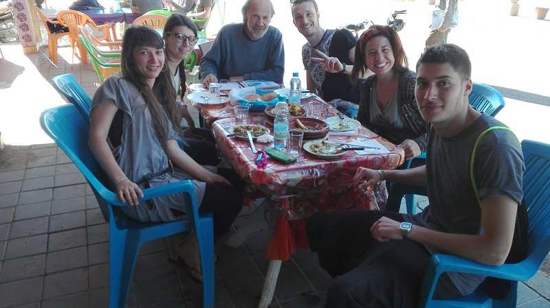 Group of five people sitting on blue plastic chairs around a table with red patterned cloth, sharing a meal at Le Gout Du Sahara Hostel.