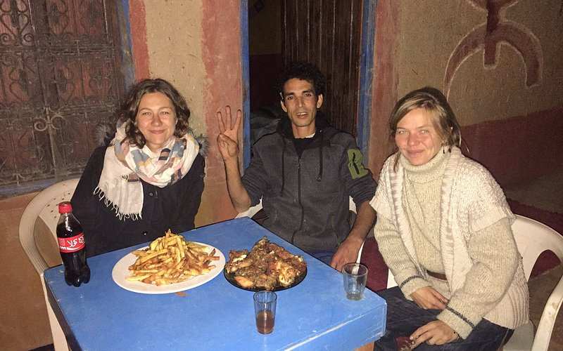 Outdoor scene at Le Gout Du Sahara Hostel showing three people sitting around a blue table with plates of fries and grilled meat and a bottle of cola.