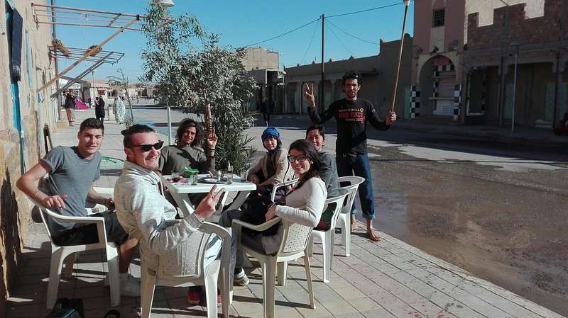 Outdoor white plastic chairs and tables with six people sitting and smiling in casual attire at Le Gout Du Sahara Hostel.