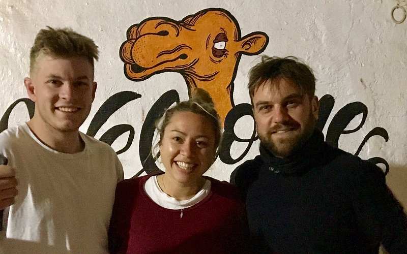 Three smiling people standing in front of a white wall with an orange camel illustration and the word welcome painted, representing Le Gout Du Sahara Hostel.