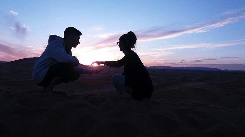 Silhouettes of two people squatting and reaching out toward each other against a colorful desert sunset sky, illustrating the serene ambiance of Le Gout Du Sahara Hostel.