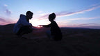 Silhouettes of two people squatting and reaching out toward each other against a colorful desert sunset sky, illustrating the serene ambiance of Le Gout Du Sahara Hostel.