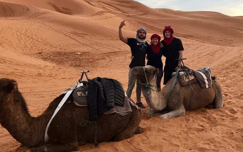 Le Gout Du Sahara Hostel guests posing with two saddled camels in orange sand dunes of Merzouga desert at sunset.