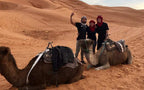 Le Gout Du Sahara Hostel guests posing with two saddled camels in orange sand dunes of Merzouga desert at sunset.
