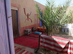 Le Gout Du Sahara Hostel outdoor seating area with red and striped rugs, colorful cushions, a palm tree, and earth-toned wall art.