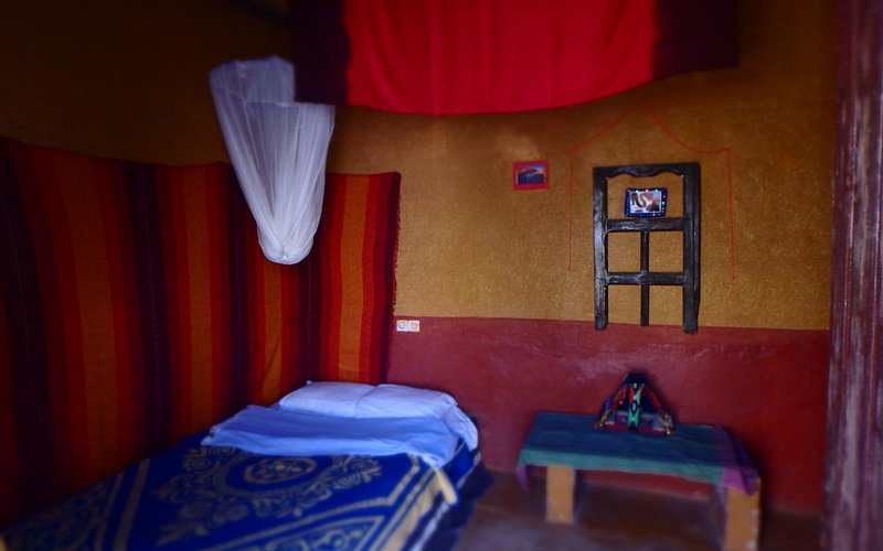 Small bedroom in Le Gout Du Sahara Hostel with warm earthy-toned walls, a single bed with blue patterned blanket, and a low table.