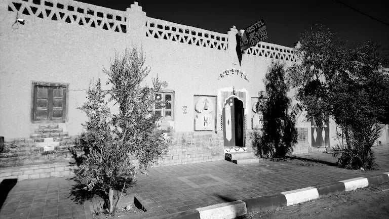 Le Gout Du Sahara Hostel shows a traditional Moroccan building with textured beige walls, arched doorways, wooden windows, and small trees in front.