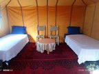 Le Gout Du Sahara Hostel bedroom with two single beds featuring white linens, blue pillows, woven chairs, and a round table on a red patterned rug.