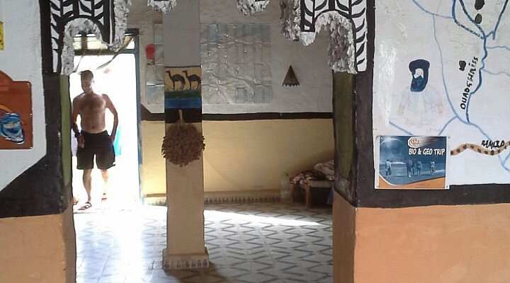 Interior view of Le Gout Du Sahara Hostel with painted white and brown walls, decorative murals, and tiled flooring, sunlight streaming in.