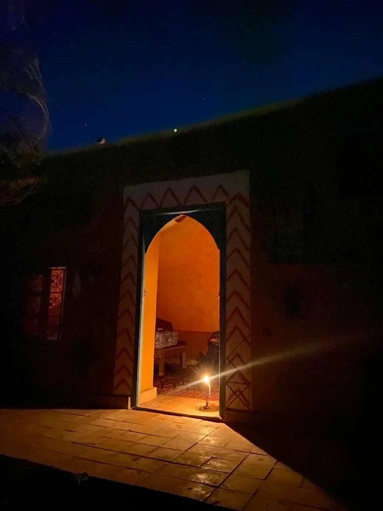 Le Gout Du Sahara Hostel entrance at night with warm orange interior light and traditional patterned archway on dark exterior wall